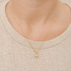 A close-up of a person's neck and upper chest wearing a delicate gold chain necklace with a small heart-shaped pendant set with clear stones, paired with a beige knit crewneck sweater.