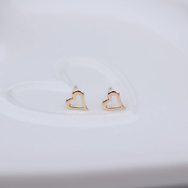 A pair of small gold heart-shaped stud earrings resting on a white ceramic dish.