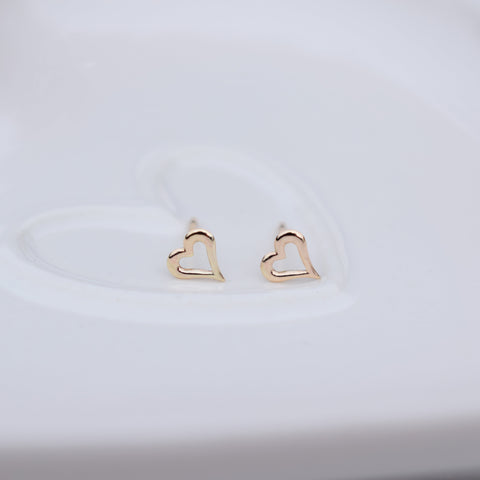 A pair of small gold heart-shaped stud earrings resting on a white ceramic dish.