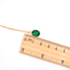 A gold chain necklace with a small oval green faceted pendant, shown next to a wooden ruler on a plain white background.