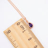 A rose gold necklace with a teardrop-shaped purple amethyst pendant, shown next to a wooden ruler for scale on a white background.