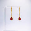 A pair of gold drop earrings with teardrop-shaped red gemstones and a small clear stone above, displayed on a clear acrylic stand against a light gray background.