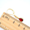 A gold leverback earring with a teardrop-shaped red gemstone, shown next to a wooden ruler for scale on a white background.