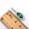 A silver-tone necklace with a marquise-shaped green gemstone pendant surrounded by clear stones, shown next to a wooden ruler for scale on a white background.
