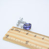 A single silver-toned stud earring with a rectangular purple gemstone, shown next to a wooden ruler on a plain white background.