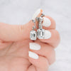 A close-up of a hand with white manicured nails holding a pair of silver-toned drop earrings with clear rectangular gemstones against a light marble background.