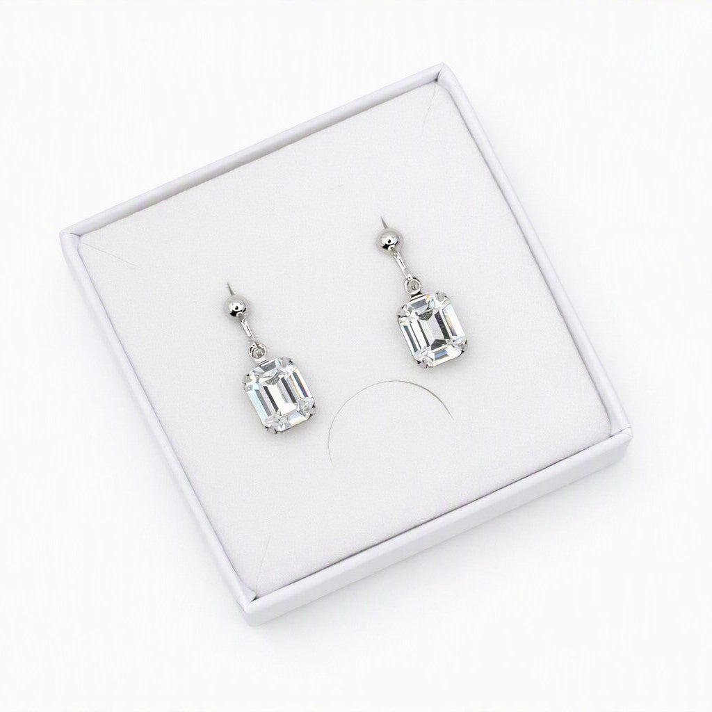 A pair of silver drop earrings with rectangular clear gemstones, presented in a white jewelry box on a white background.