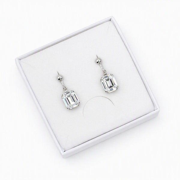A pair of silver drop earrings with rectangular clear gemstones, presented in a white jewelry box on a white background.