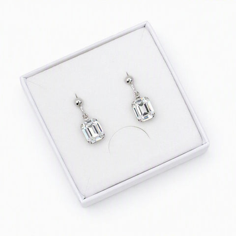 A pair of silver drop earrings with rectangular clear gemstones, presented in a white jewelry box on a white background.