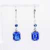 A pair of silver drop earrings with rectangular blue gemstones and small round blue accent beads, displayed on a clear acrylic stand against a white background.