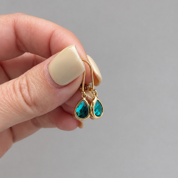 A close-up of a hand with beige manicured nails holding a pair of gold drop earrings with teardrop-shaped green gemstones against a solid gray background.