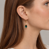 A close-up side profile of a woman with long brown hair wearing a gold dangle earring with a dark green faceted gemstone and a small clear accent stone.