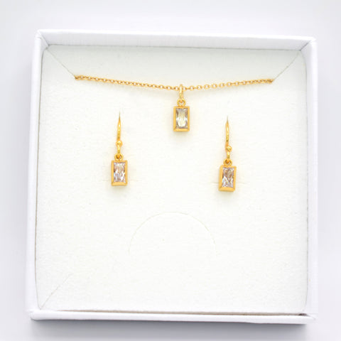Gold jewelry set including a delicate chain necklace with a rectangular pendant and matching drop earrings, displayed inside a white square gift box on a white background.