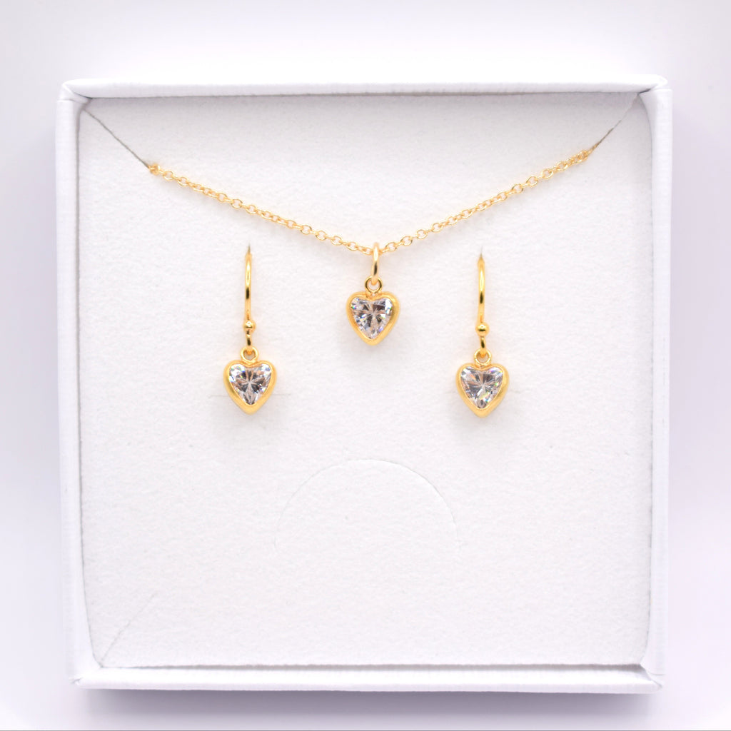 A gold jewelry set featuring a delicate chain necklace and two matching drop earrings, each with a small heart-shaped clear gemstone in a gold setting, presented inside an open white gift box.