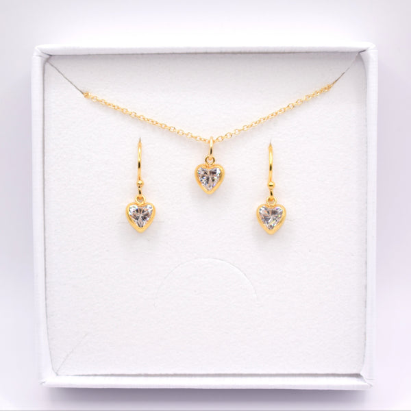 A gold jewelry set featuring a delicate chain necklace and two matching drop earrings, each with a small heart-shaped clear gemstone in a gold setting, presented inside an open white gift box.