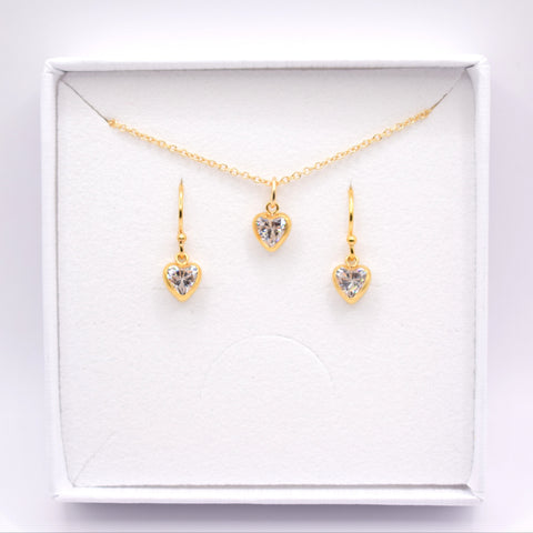 A gold jewelry set featuring a delicate chain necklace and two matching drop earrings, each with a small heart-shaped clear gemstone in a gold setting, presented inside an open white gift box.