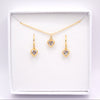 A gold jewelry set featuring a delicate chain necklace and two matching drop earrings, each with a small heart-shaped clear gemstone in a gold setting, presented inside an open white gift box.