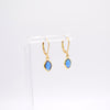 A pair of gold hoop earrings with marquise-cut blue gemstones, displayed on a clear acrylic stand against a white background.