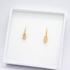 A pair of gold drop earrings with rectangular clear gemstones, displayed on a white jewelry box against a white background.