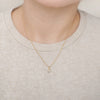 A close-up of a model wearing a delicate gold chain necklace with a small, clear rectangle-shaped crystal pendant. The model is wearing a light beige crewneck sweater against a soft, neutral background.